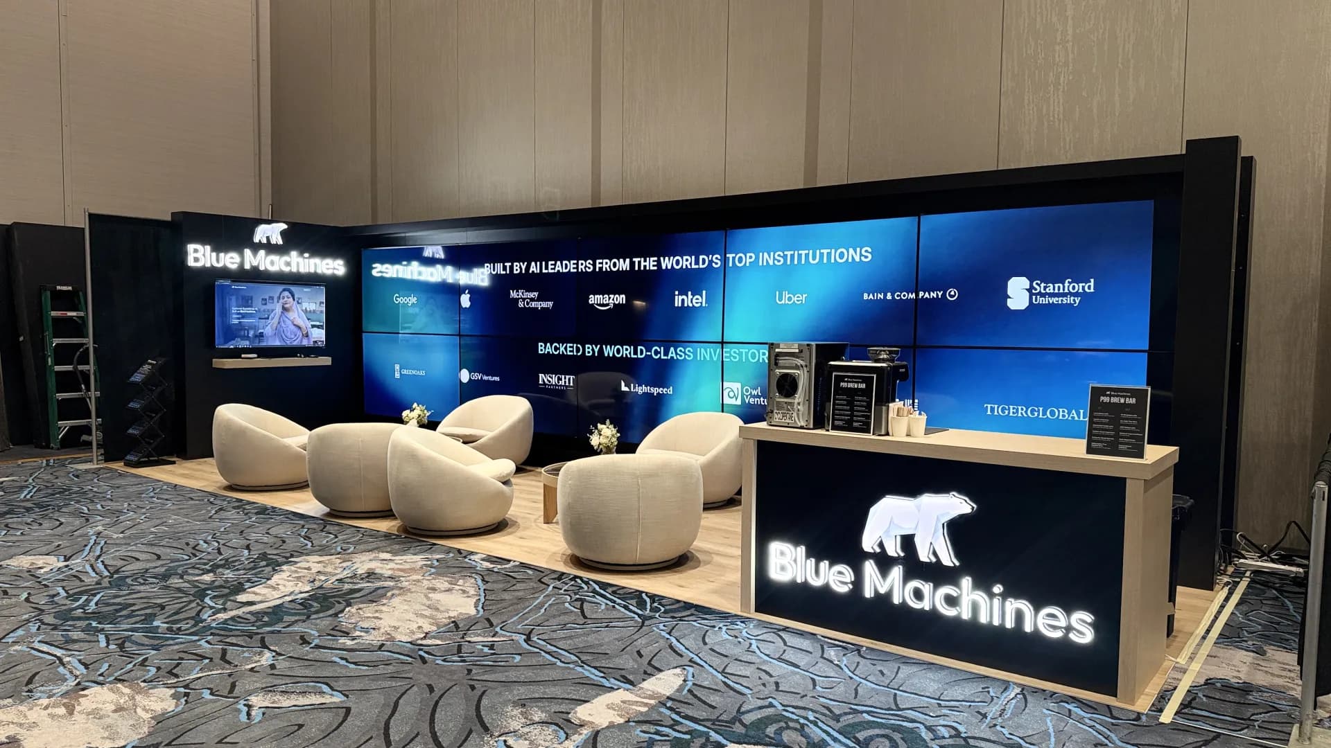 Wide view of the Blue Machines island booth with large-format partner-logo graphics on the video wall